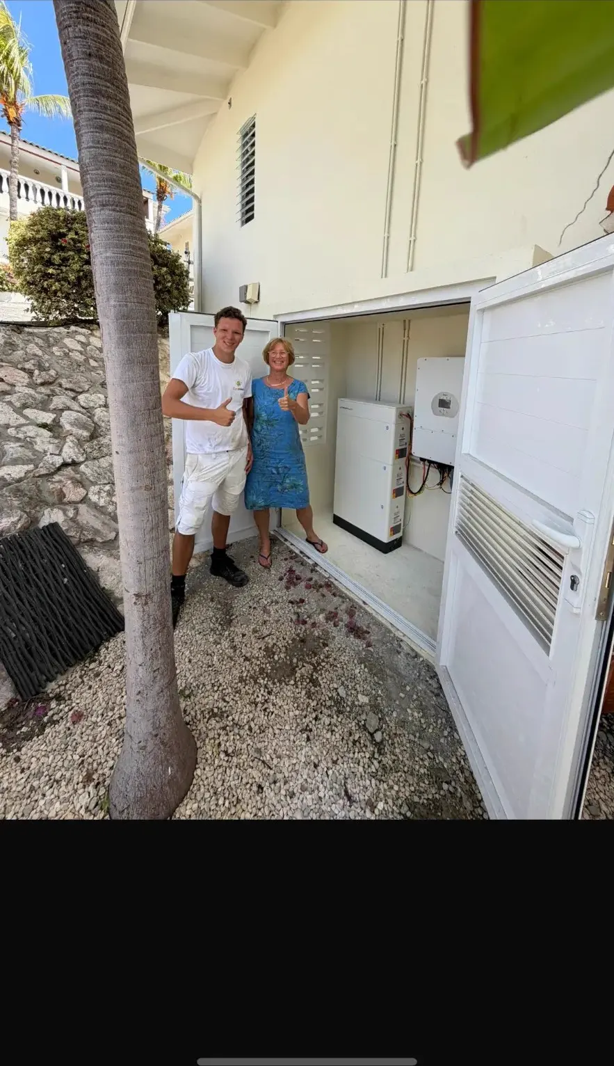 ZonZeker-monteur en tevreden klant bij een stapelbare thuisbatterij met omvormer in een buitenberging op Curaçao