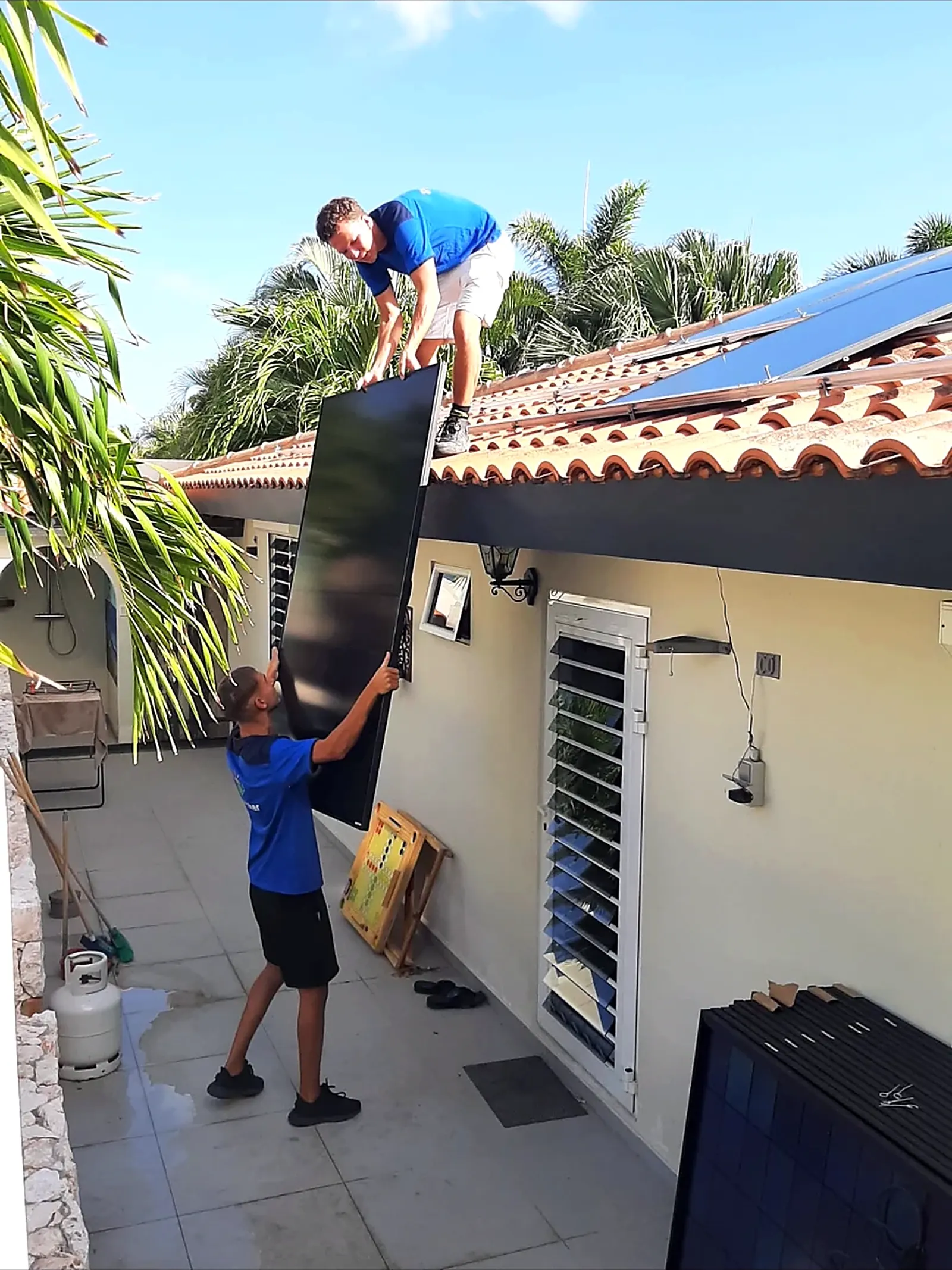 Twee ZonZeker-monteurs geven een zwart zonnepaneel aan naar het pannendak van een woning op Curaçao