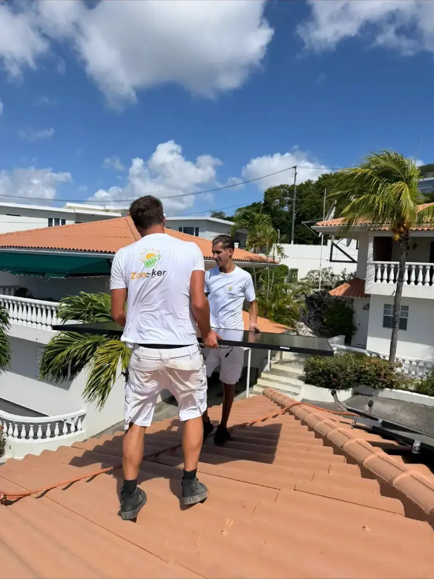 Twee ZonZeker-monteurs dragen samen een zonnepaneel over een dak in Curaçao